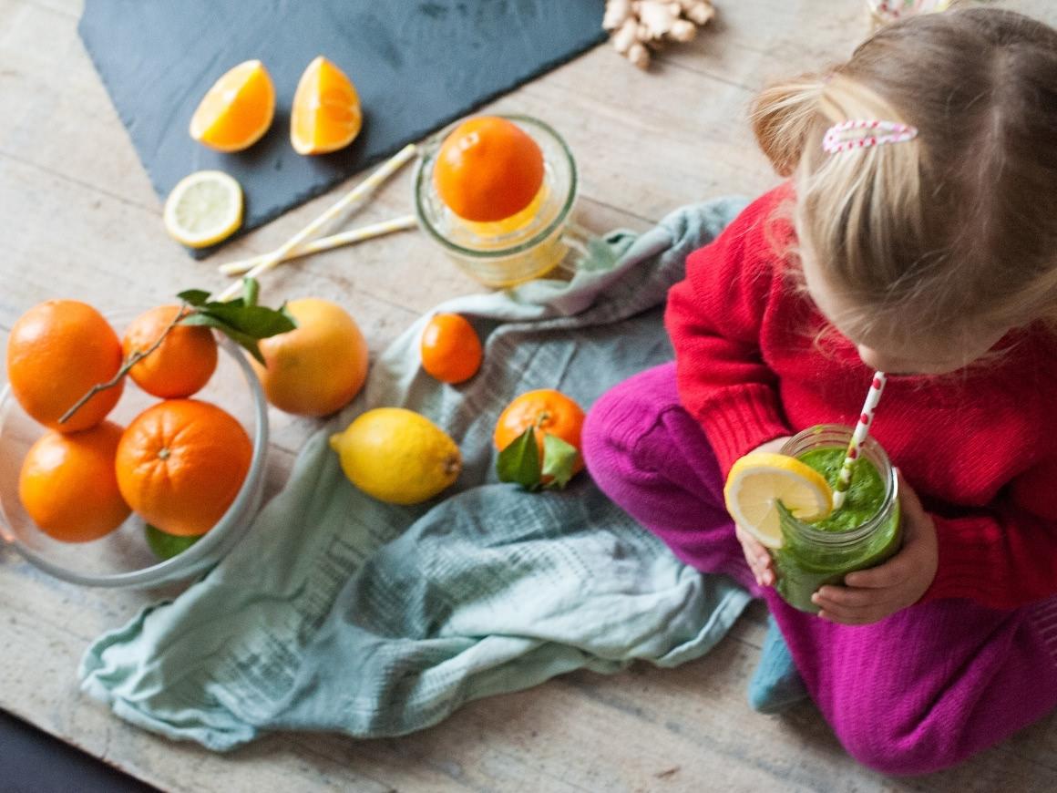 little girl sitting on a table, drinking a green smoothie which is decorated with a slices of lemon. On the table are oranges, tangerines, lemons.